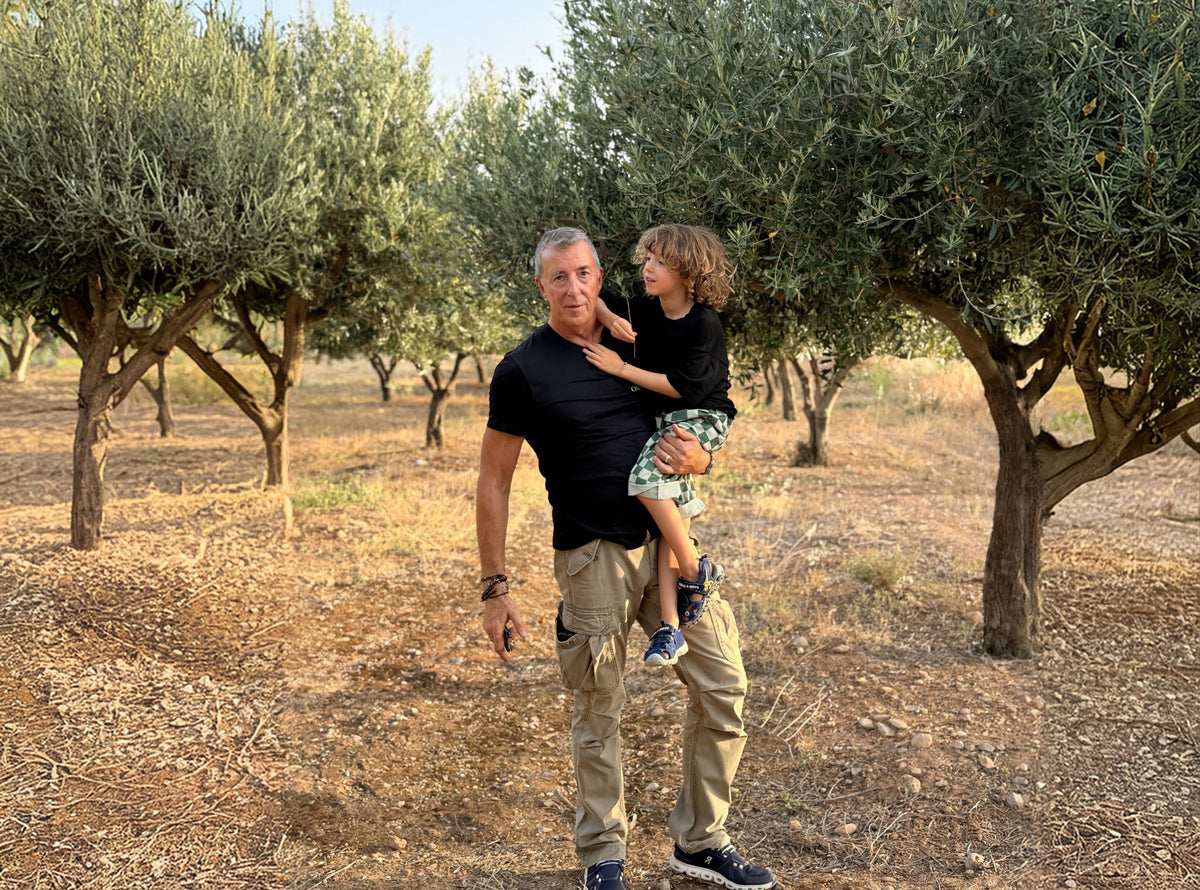 A man carrying a young child in an olive grove with lush green trees.