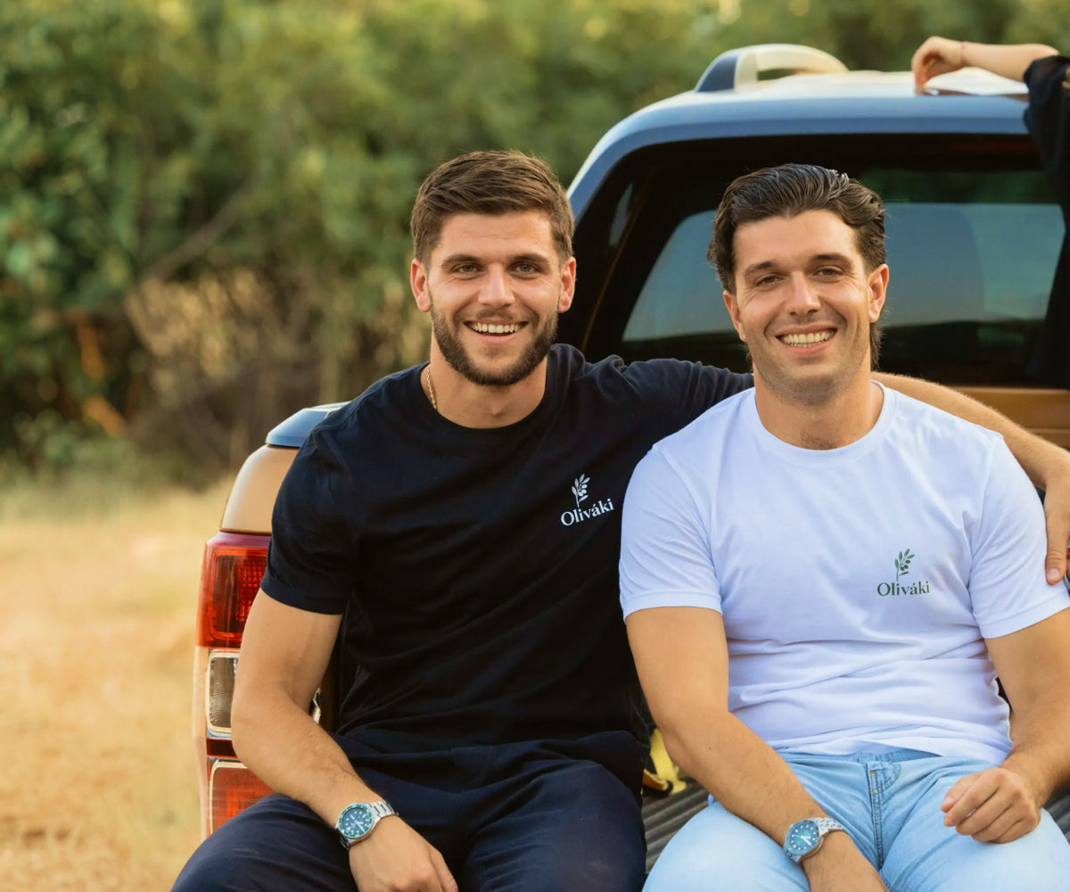 Two smiling men wearing Oliváki branded t-shirts sitting in the back of a pickup truck outdoors.