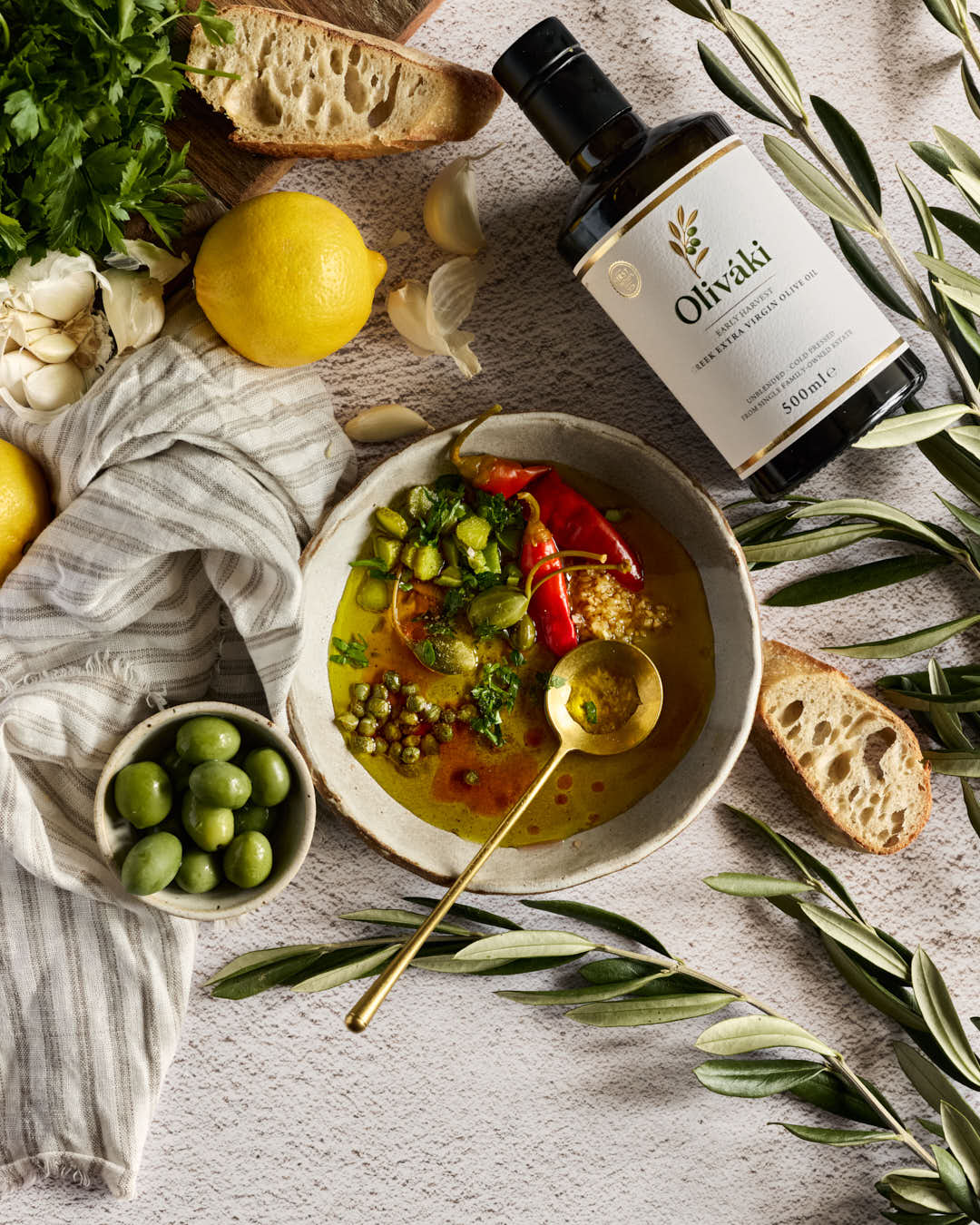 A styled overhead flat-lay of a bowl of olive oil soup with a gold spoon, surrounded by ingredients including a bottle of Olivaki olive oil, bread, garlic, parsley, green olives, and olive branches on a textured light gray surface.