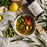 A styled overhead flat-lay of a bowl of olive oil soup with a gold spoon, surrounded by ingredients including a bottle of Olivaki olive oil, bread, garlic, parsley, green olives, and olive branches on a textured light gray surface.