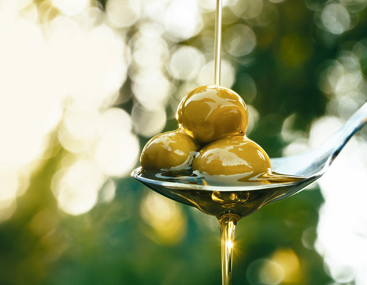 Green olives drizzled with golden extra virgin olive oil on a silver spoon with bokeh background.