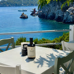 A bottle of Oliváki Greek Extra Virgin Olive Oil on a white table overlooking a blue Mediterranean sea.