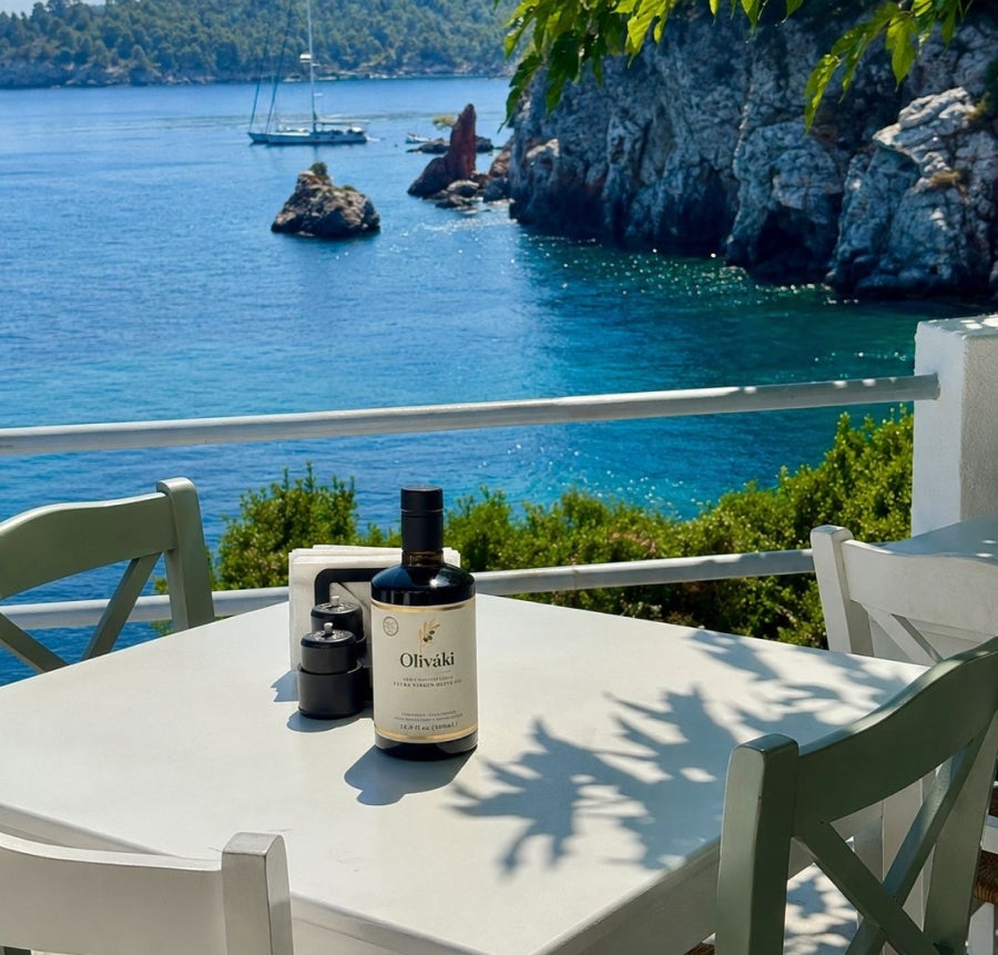 A bottle of Oliváki Greek Extra Virgin Olive Oil on a white table overlooking a blue Mediterranean sea.
