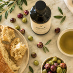 Oliváki Greek extra virgin olive oil bottle with olives and crusty bread on marble.