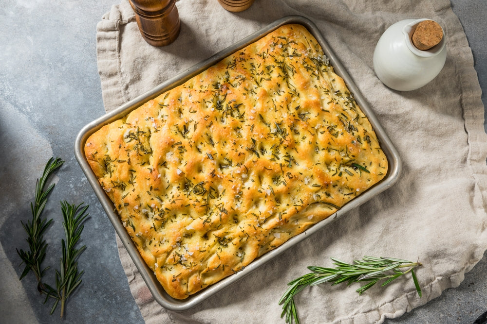 Freshly baked rosemary focaccia bread in a rectangular metal baking pan on a linen cloth.