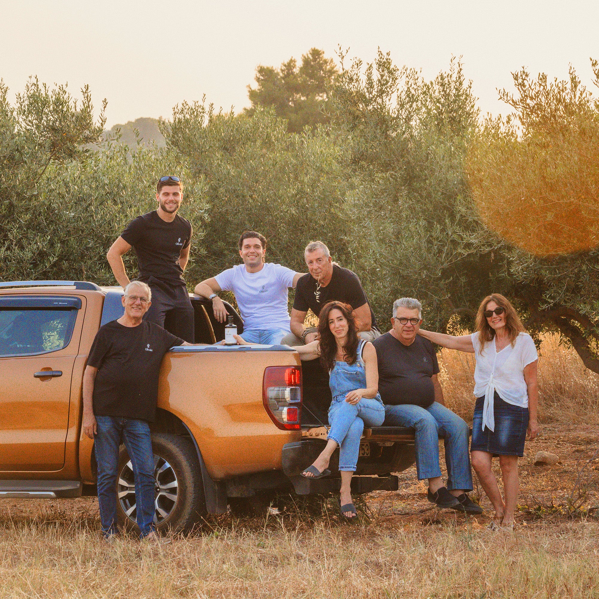 A smiling group of people posing together by an orange pickup truck in an olive grove.